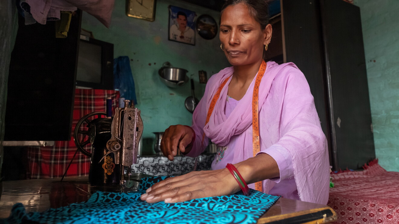 A woman sits at a table with a sewing machine. She has a measuring tape draped around her neck and uses her right hand to position a patterned fabric.