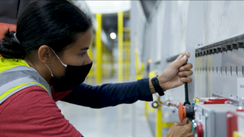 A woman wearing a mask and safety vest pulls down on a lever.