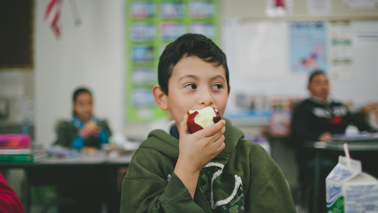 A boy in a classroom is eating an apple. Behind him, other students are seated at desks.