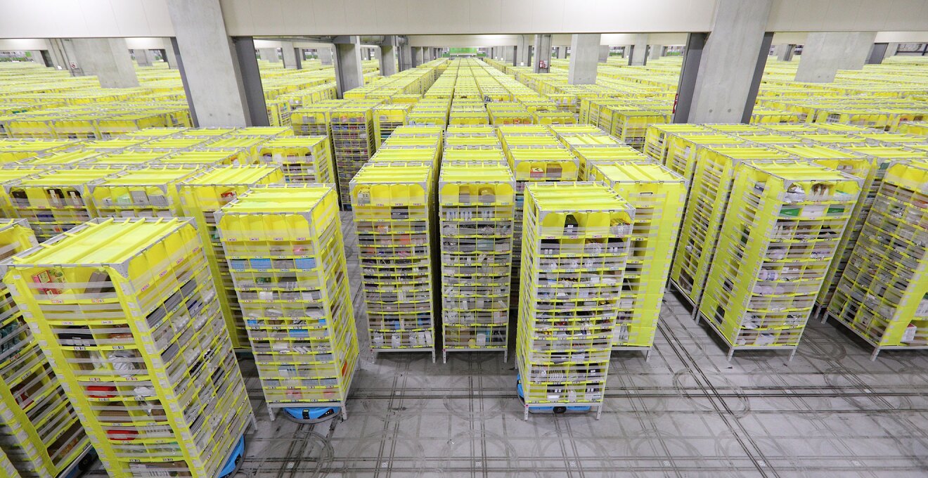グレーの広大な物流センター内。整然と並ぶ黄色い収納棚の列が、視界のいちばん奥まで続いている。