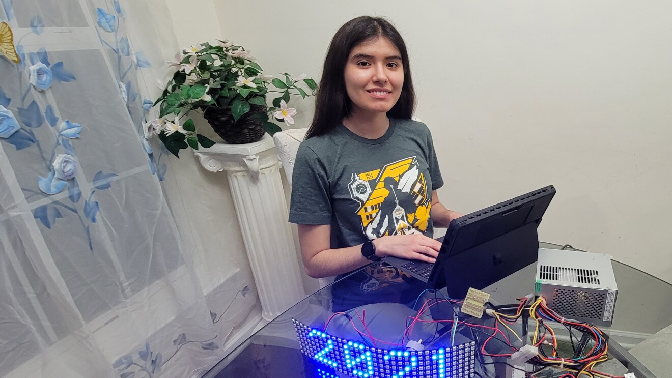 An image of a young woman sitting at a dining room table smiling for a photo. In front of her is a laptop hooked up to wires and an illuminated sign that says "2021."