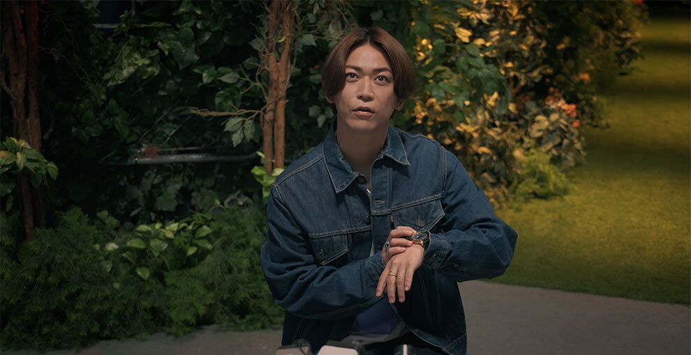 緑の植栽と人工芝の薄暗い背景に、インディゴブルーのデニムジャケットを着た人物が立っている。上方を見上げるポーズで、胸の高さで左手の腕時計に右手を添えている。