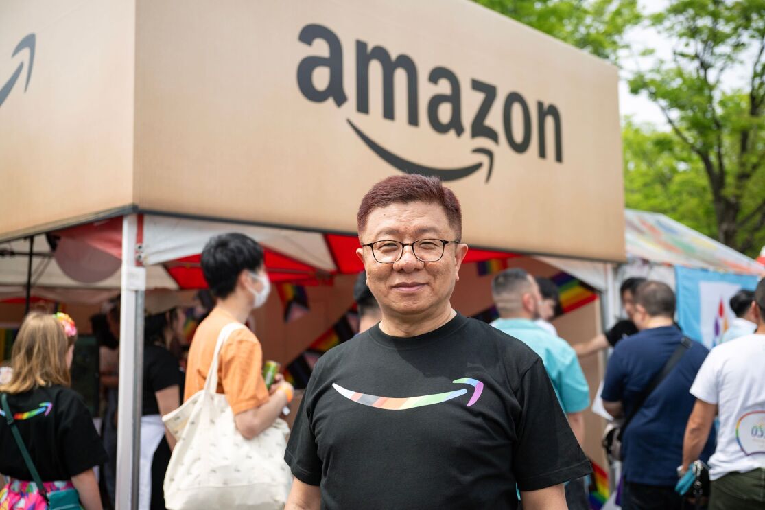 Amazonと書かれたブースの前に立つメガネをかけたアジア系の男性。黒いTシャツを着ている