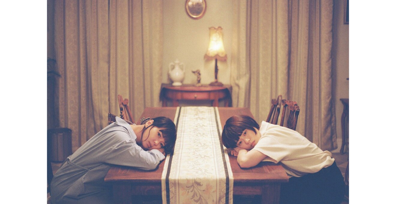 長方形のテーブルをはさんで左右に女性が1人ずつ座り、共にテーブルに突っ伏して腕にのせた顔をこちらに向けている。2人の頭の間にはテーブルランナー。