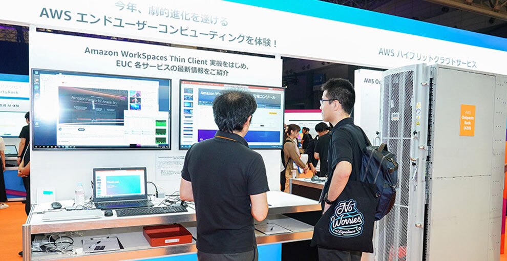 展示会の会場内、AWSハイブリットクラウドサービスのブース前に立つ2人の人物。ブースの上部には「今年、劇的進化を遂げるAWSエンドユーザーコンピューティングを体験!」とある。2人の視線の先にはモニターが2枚あり、EUCのサービスが紹介されている。デスク部分には開いたノートPCとキーボード。