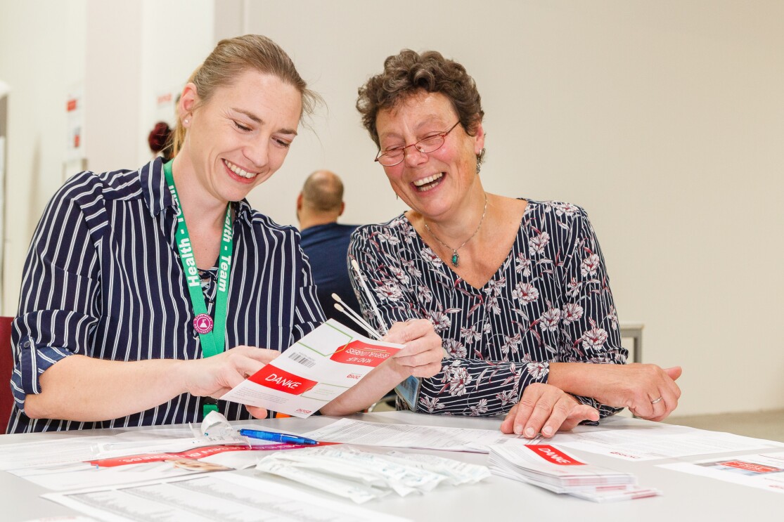 Zwei lächelnde Mitarbeiterinnen sitzen an einem Tisch, vor ihnen DKMS-Registrierkarten und Wattestäbchen
