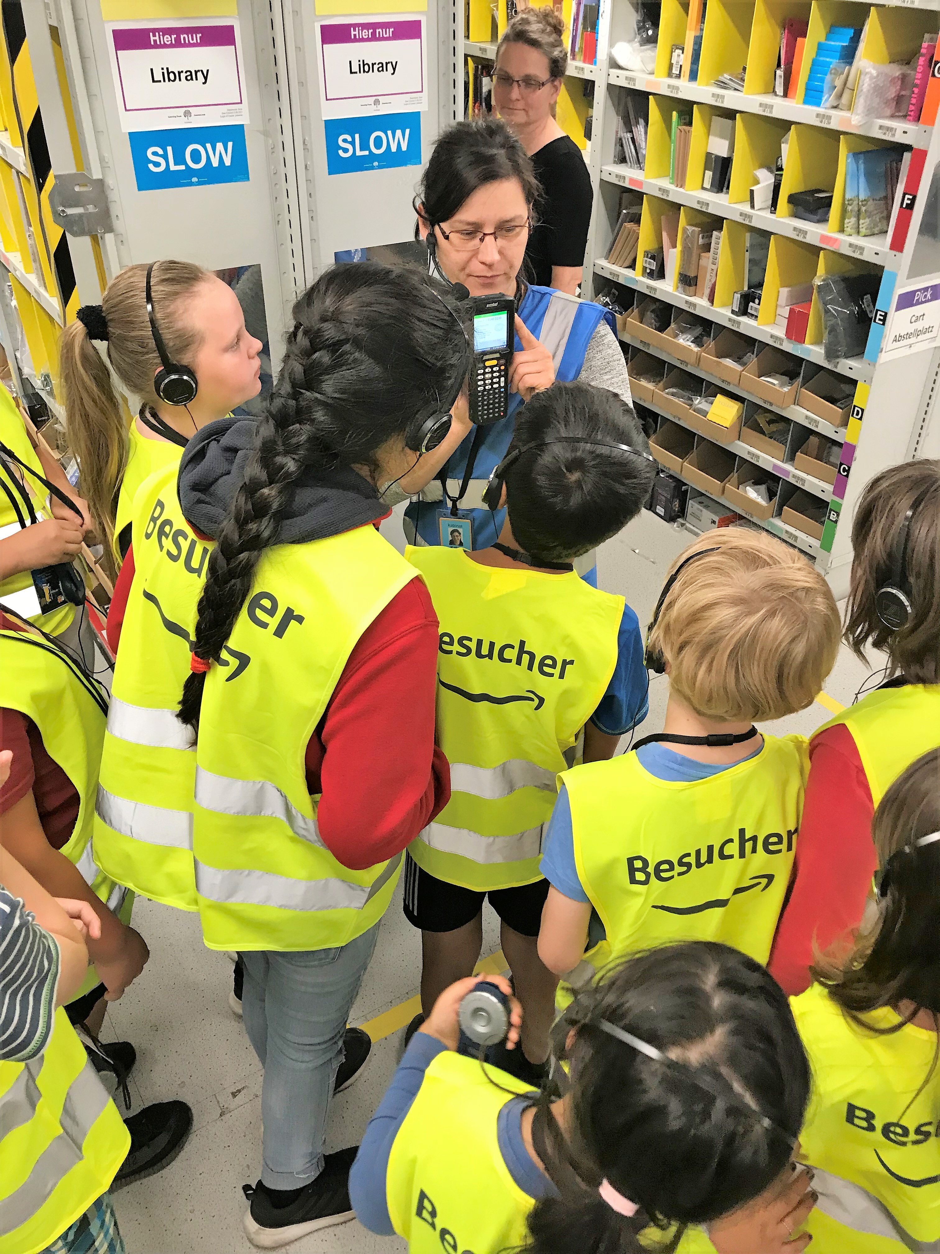 Eine dunkelhaarige Amazon Mitarbeiter mit blauer Sicherheitsweste ist von Kindern in gelten Sicherheitswesten mit Aufdruck "Besucher" umringt: Sie zeigt auf den Scanner, dessen Funktionsweise sie gerade erklärt.