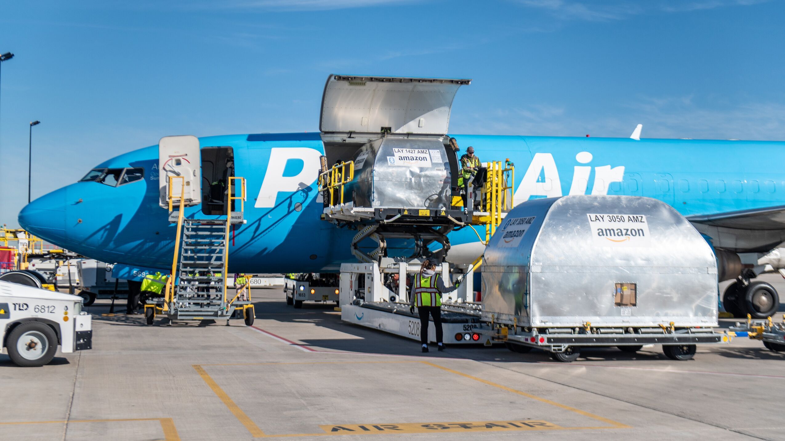 An image of a light blue Amazon Air plane. The door on the side of the plane is open and employees are loading a large, silver container inside the plane. There are several lifts and stair sets attached to the open doors on the plane for employees to access the inside and load it with containers carrying Amazon packages.