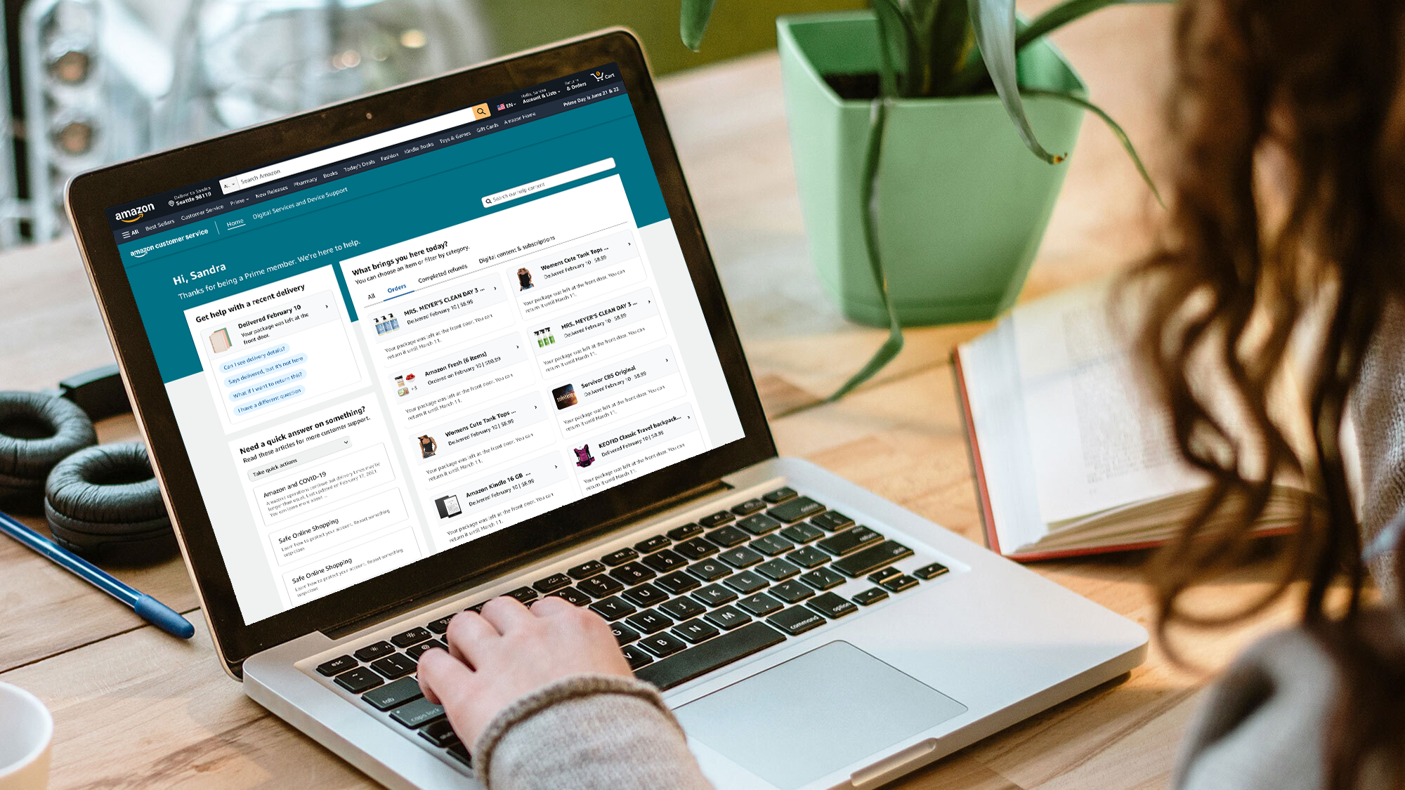 Woman on sitting at a table working on a laptop. On the screen of the laptop is the Amazon Customer Service homepage.
