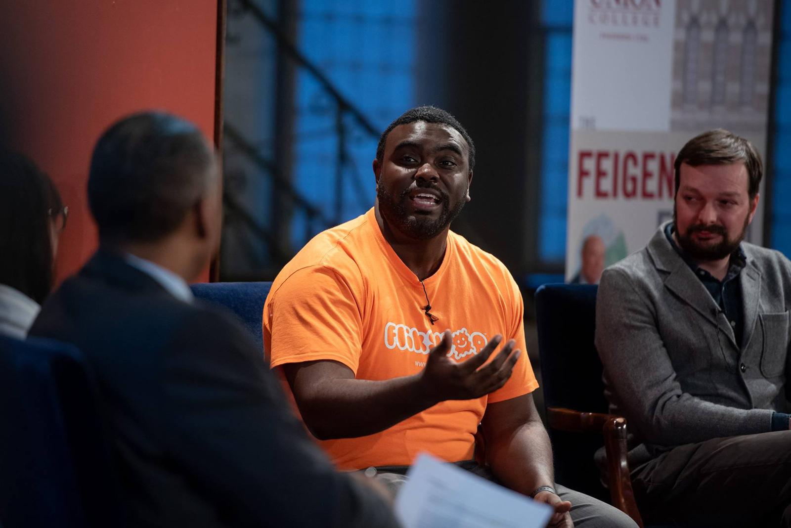 An image of a man speaking to a panel of people. He is wearing an orange shirt that says "Flikshop."