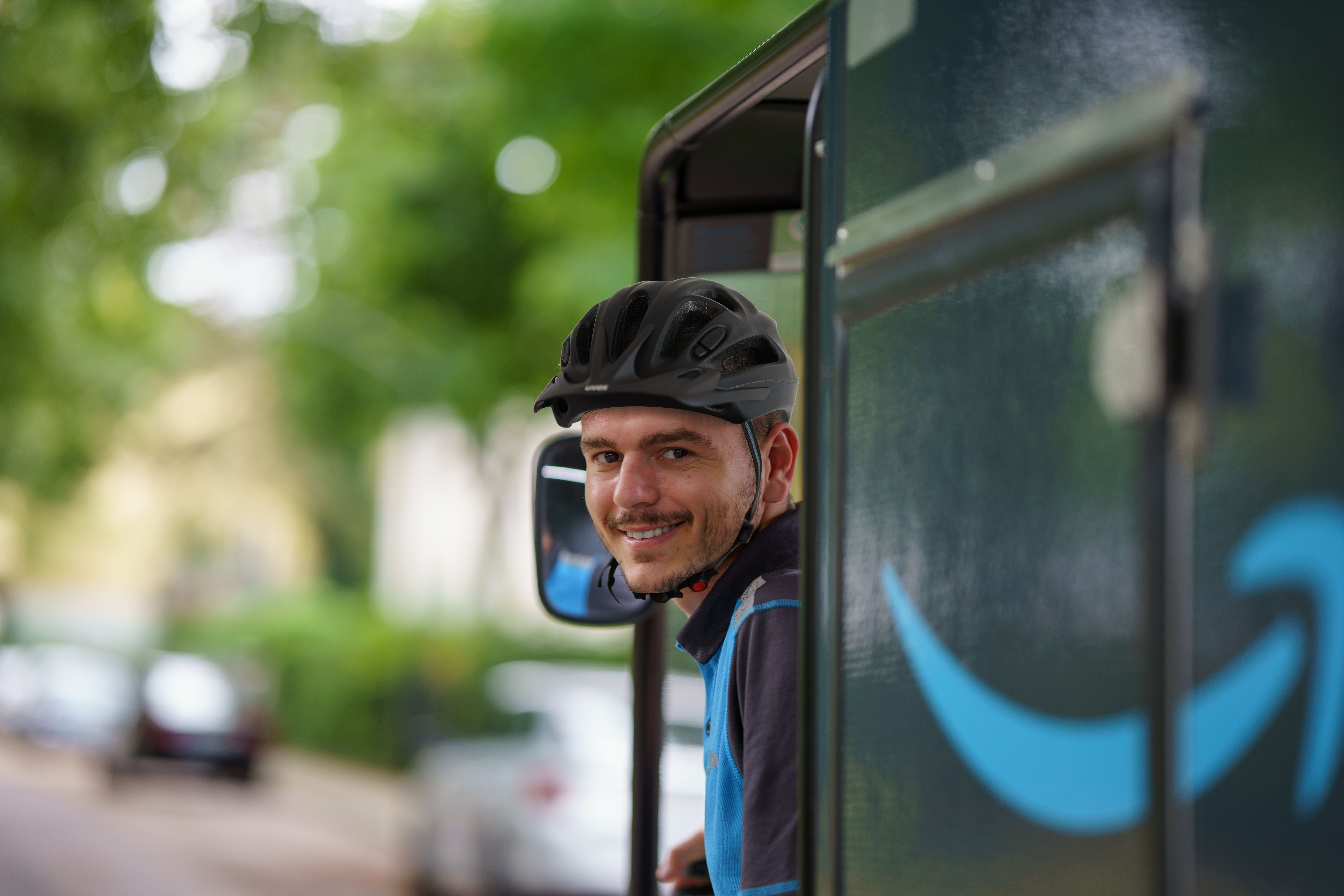 Ein junger Amazon E-Cargo-Bike-Fahrer mit schwarzem Helm und blauem Shirt auf dem E-Cargo-Bike. Er blickt sich um und lächelt in die Kamera. Verschwommen sieht man das seitlich angebrachte Amazon-Logo.