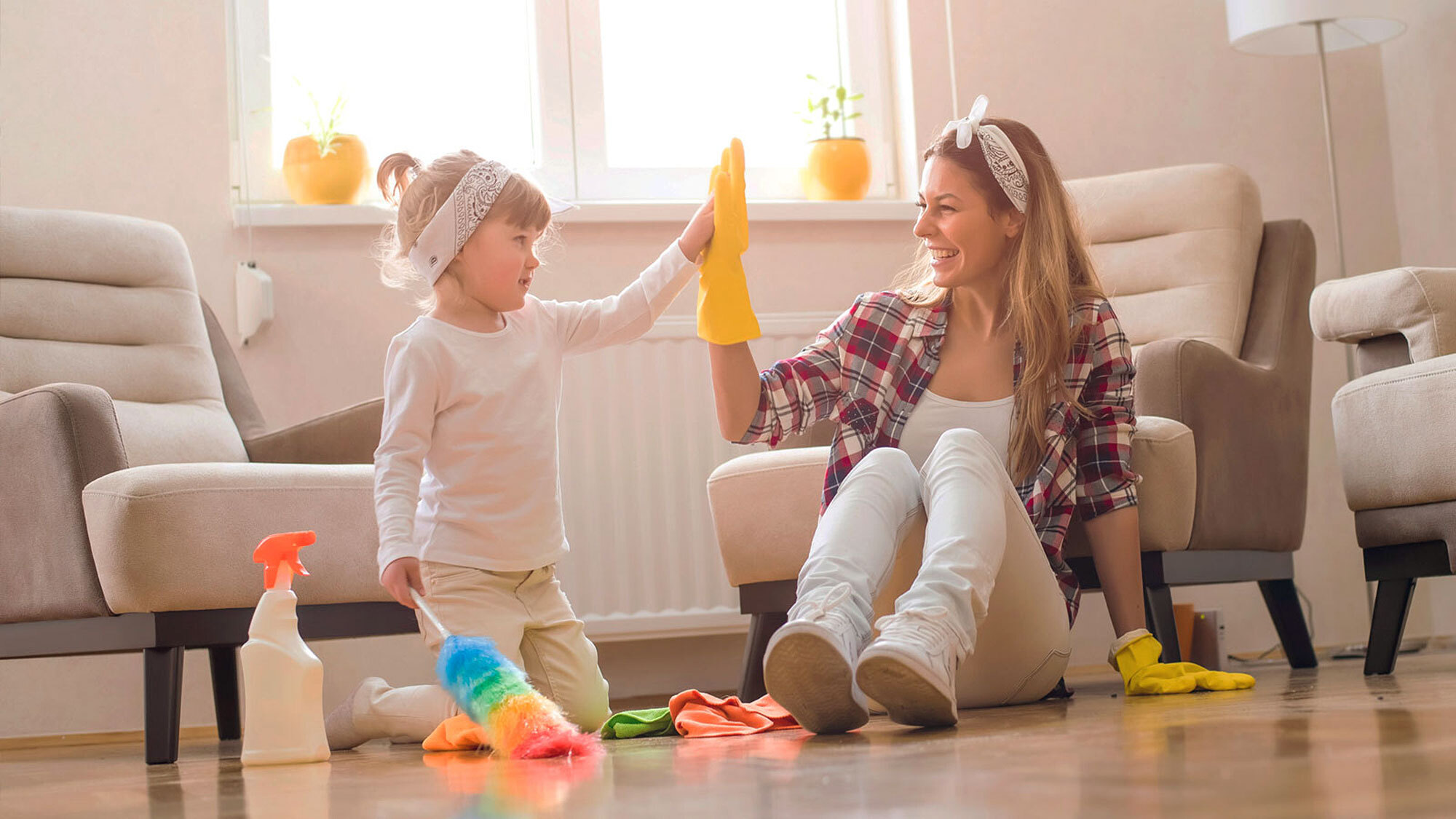 A mom smiles and gives her daughter a high five as they sit on the floor next to cleaning supplies in a living room.