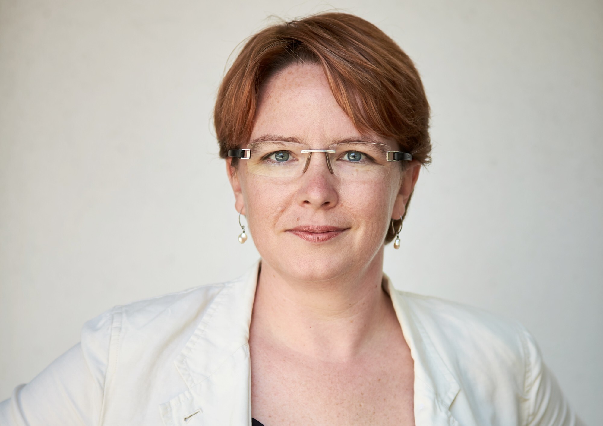 A woman smiles for a headshot photo in front of a white background while wearing a white blazer and glasses.