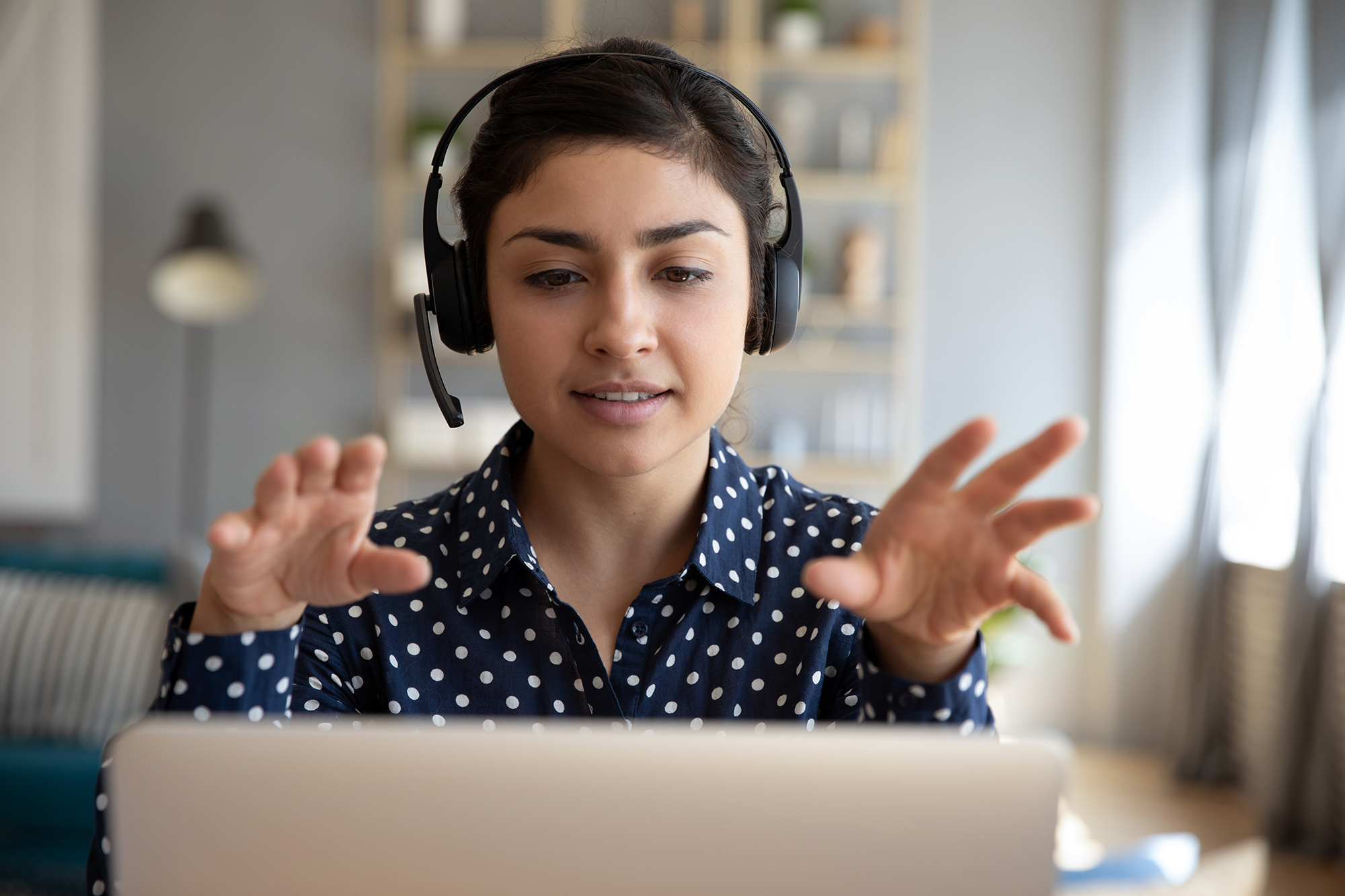 A woman wearing a button down shirt and headset looks into her laptop screen while gesturing with her hands. 