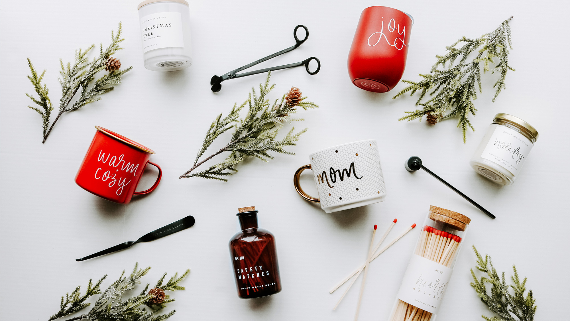 Small gifts spread out on a white background and among evergreen tree branches. Gifts include mugs, candles, and candlestick scissors.