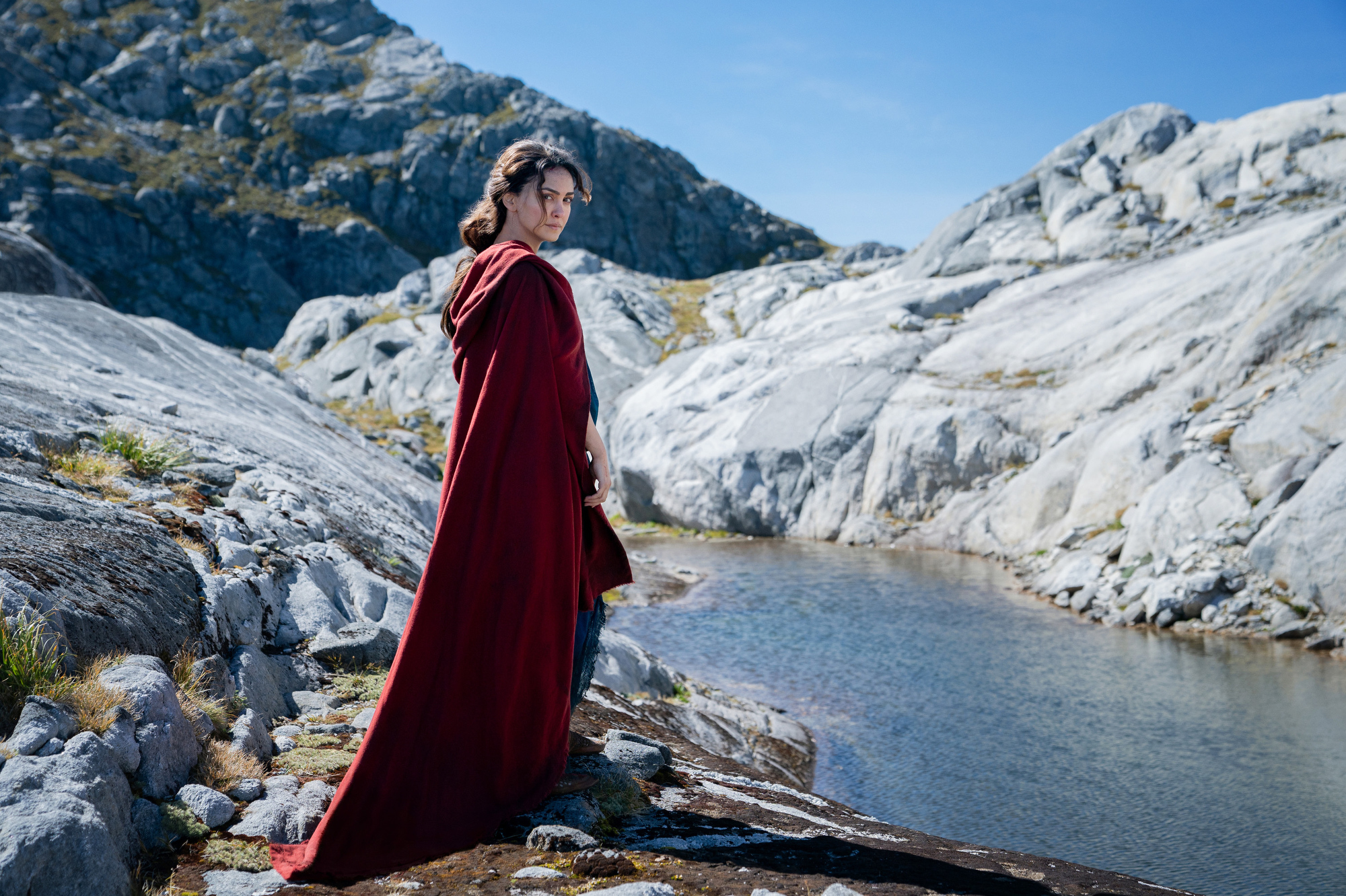 A female character wears a red cape in a rocky area while standing above a river.