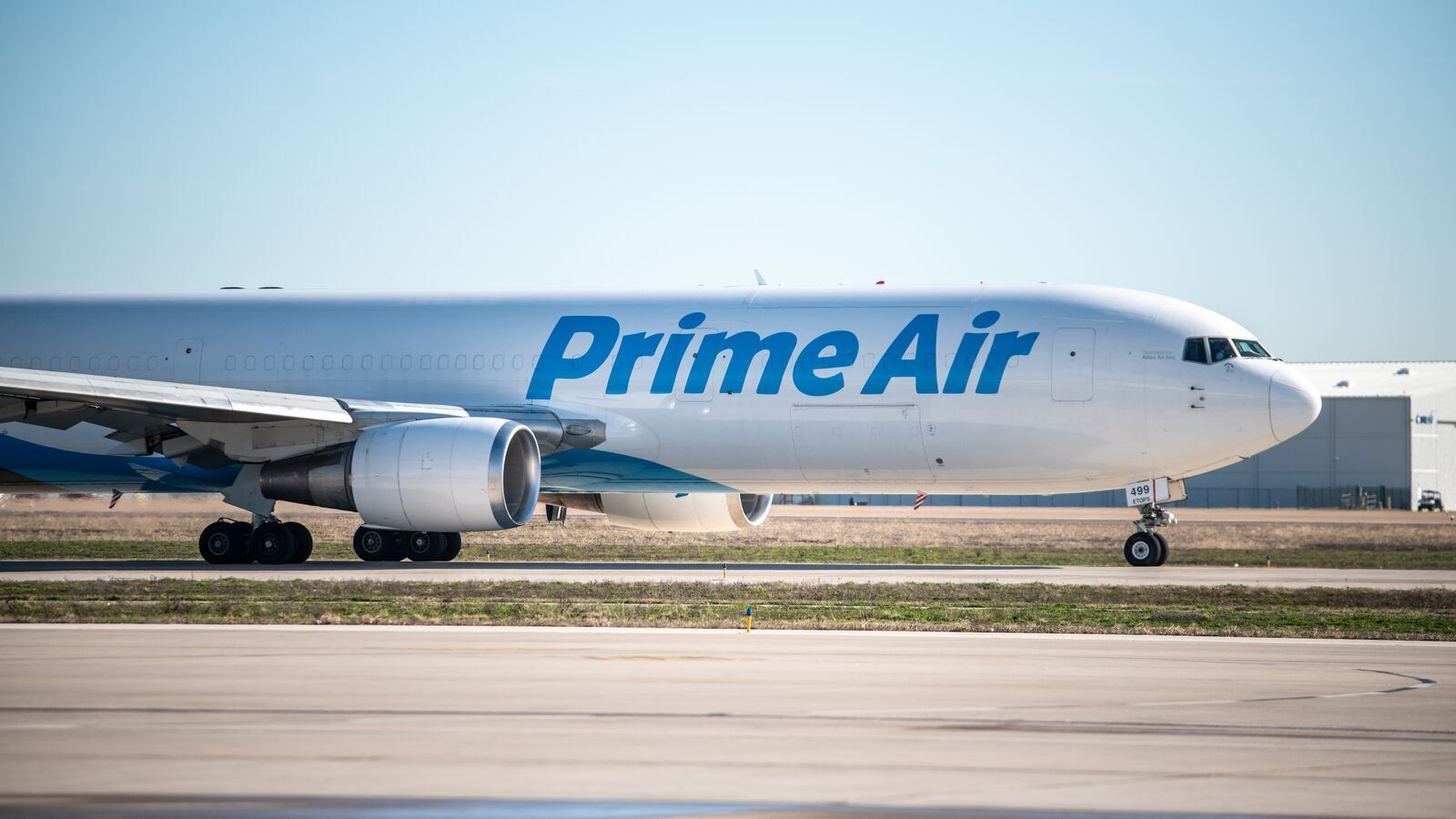 An image of an Amazon Air plan getting ready for takeoff. It is sitting on the runway in a large field. The plane is white with blue writing that says "Prime Air."