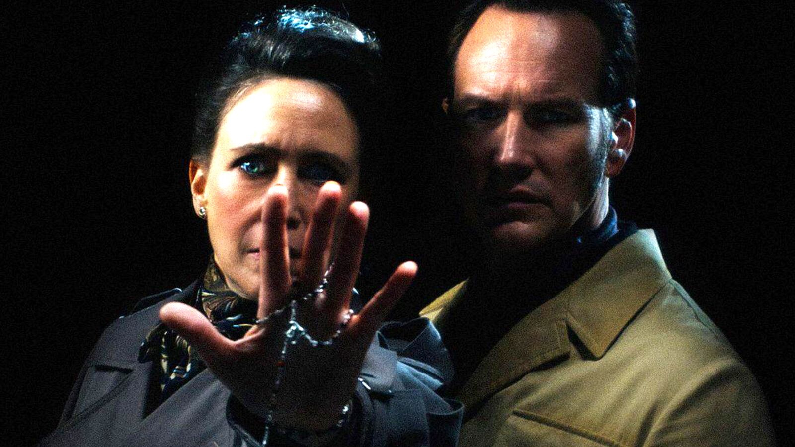 The Conjuring movie: Two intense figures in dark setting, woman's hand reaching toward viewer

