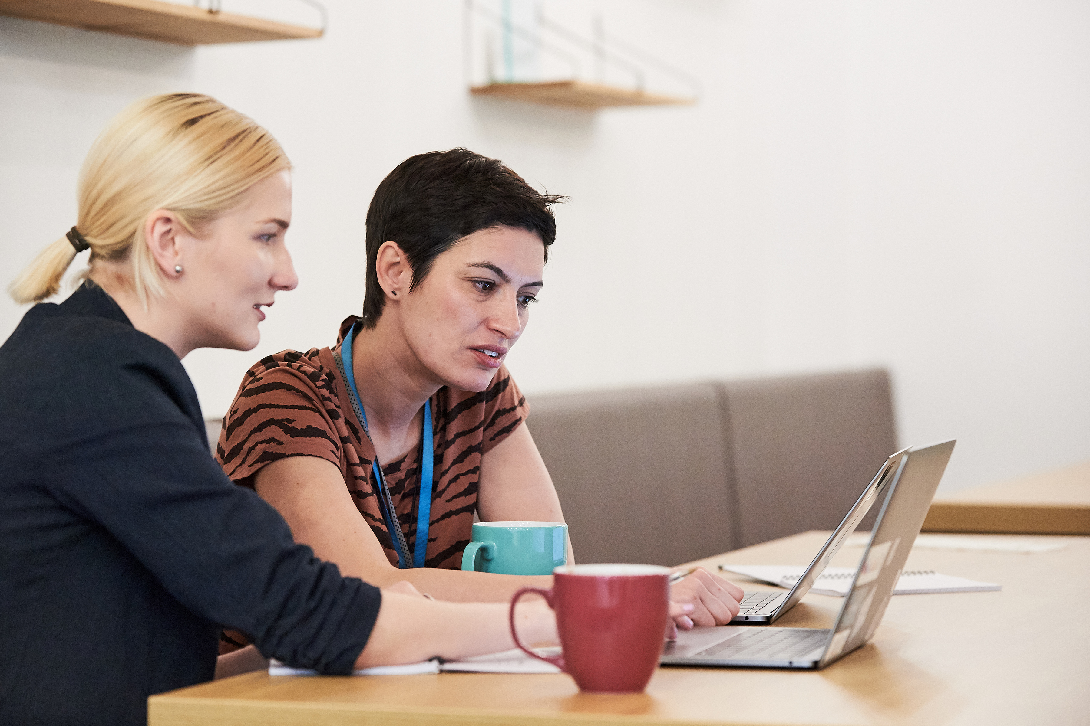 Zwei Frauen sitzen an einem Holztisch und schauen in einen Laptop. Auf dem Tisch stehen zwei Kaffeetassen.