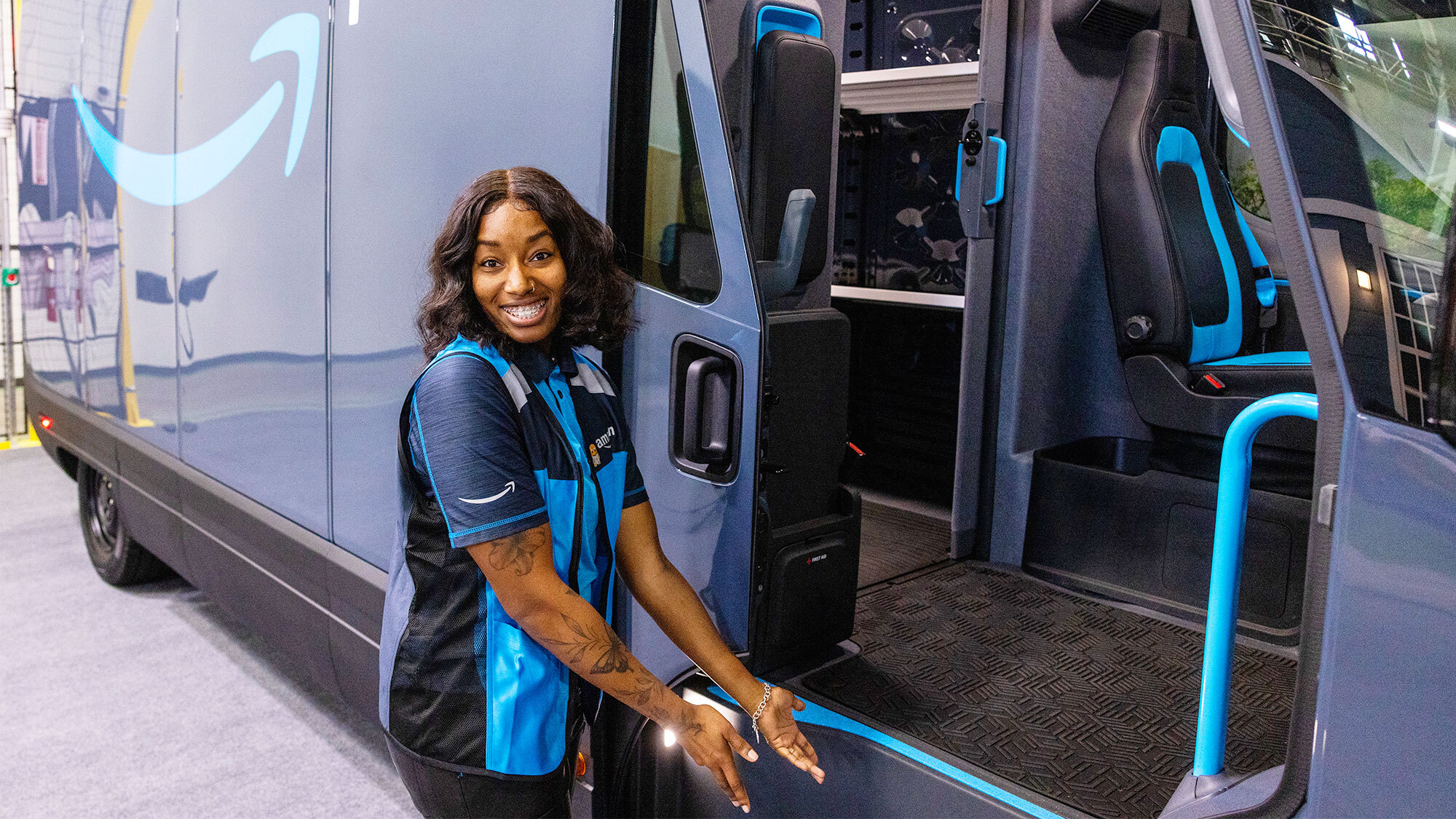 A photo of Brandi Monroe, a delivery driver for Kangaroo Direct, an Amazon Delivery Service Partner in the Baltimore, Maryland area. Brandi is standing at the front of the entrance door to an Amazon delivery van from Rivian, with arms open to the staircase.