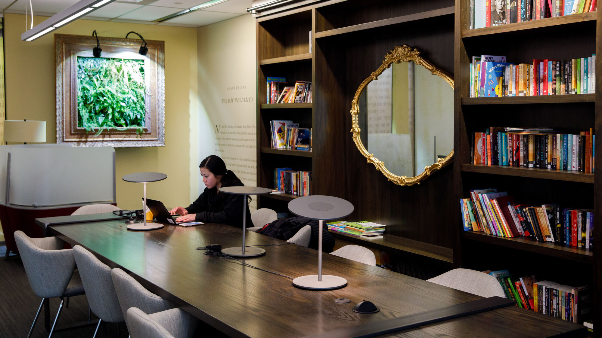 An image of a woman working on her laptop while sitting at a large table inside an Amazon office in Seattle. There is a large bookshelf behind her and a "Harry Potter" quote on the wall.