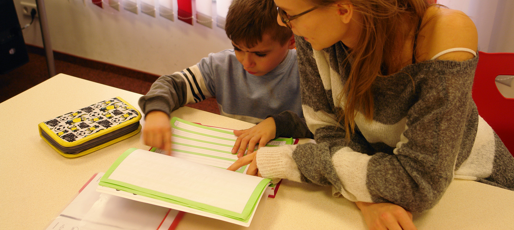 Ein kleiner Junge und eine junge Frau sitzen gemeinsam an einem Tisch und schauen auf ein Schulheft.