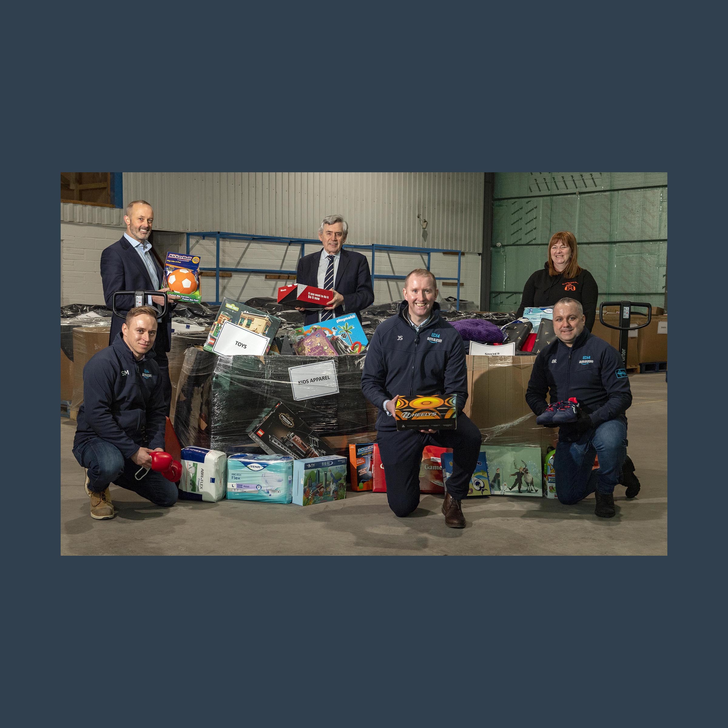 Six individuals pose together in a warehouse setting with products to be donated to those in need