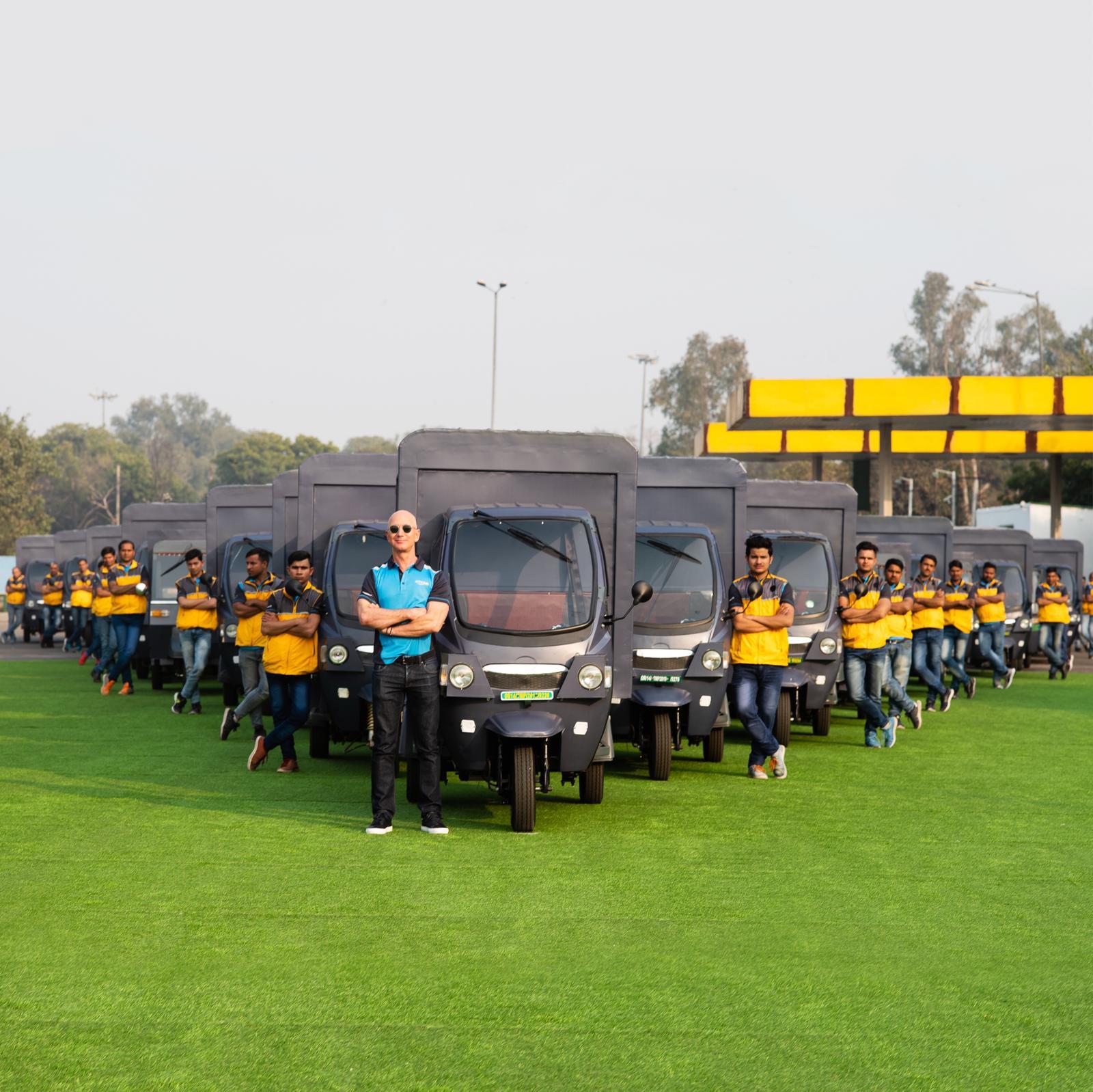 Jeff Bezos stands next to an electric rickshaw, behind him stand several other Amazon delivery drivers who also stand next to electric rickshaws.