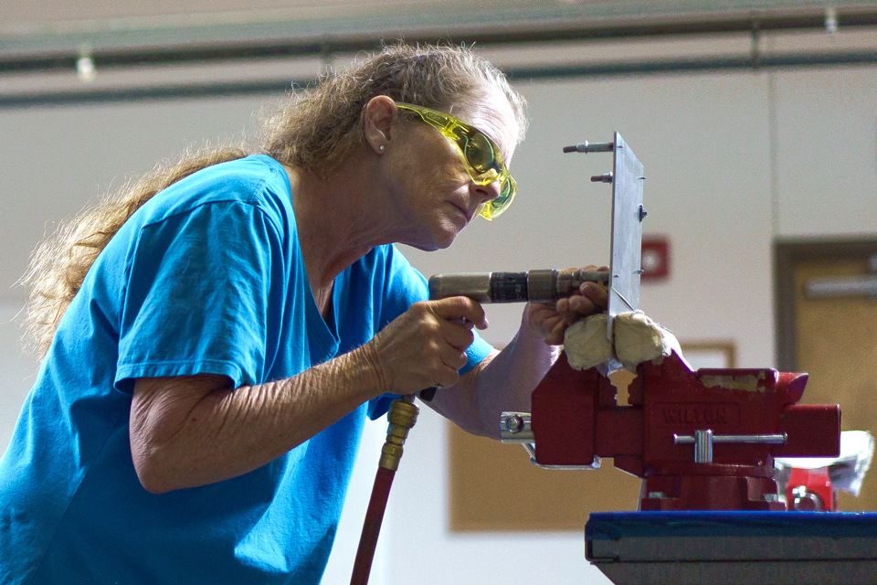 A woman in a blue T-shirt and safety glasses applies a hydraulic tool to a sheet of metal and leans toward a workbench vise.