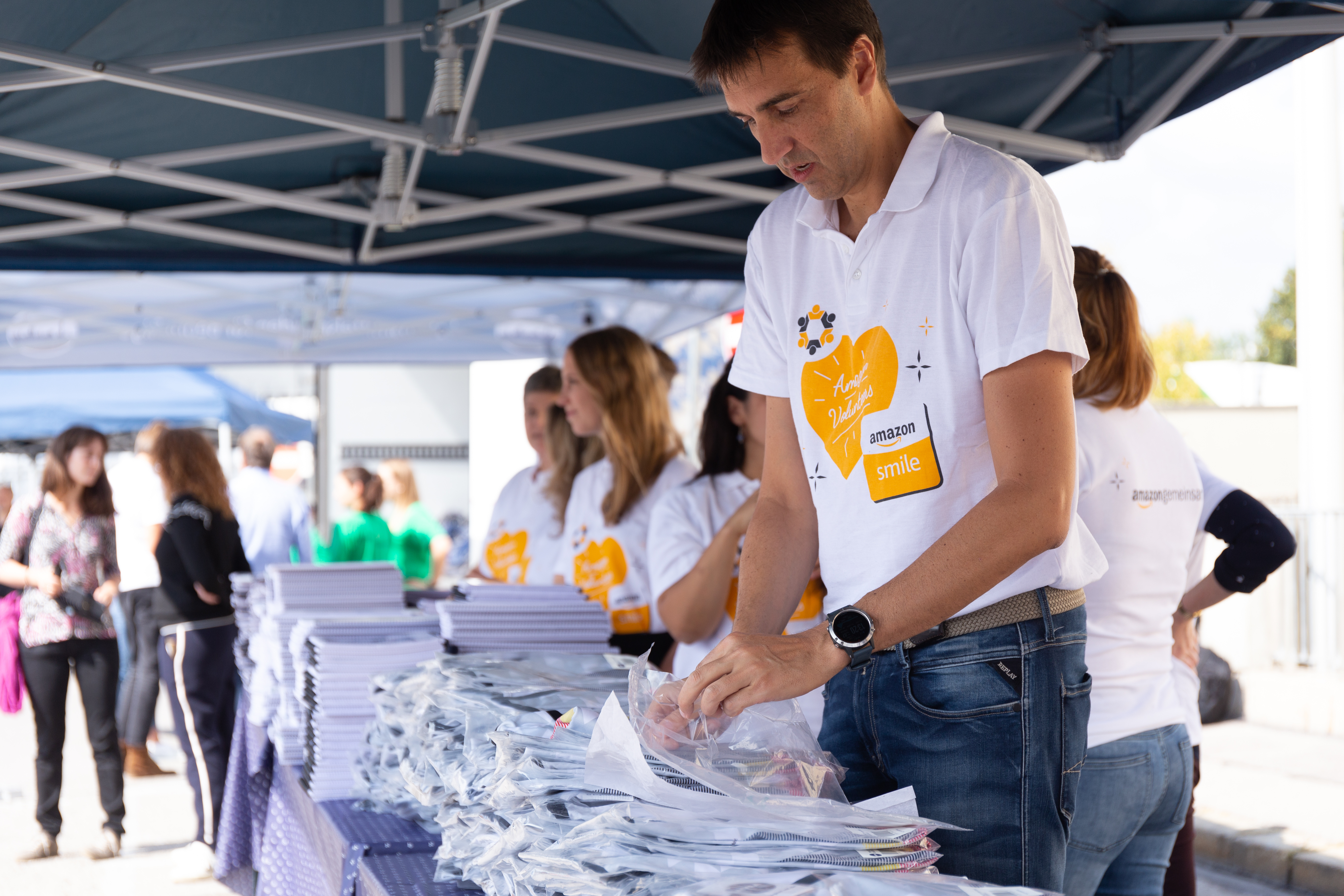 Amazon Mitarbeiter helfen beim Verteilen von Schulmaterial, wie Bücher, Hefte, Mappen. Alle tragen das AmazonSmile T-Shirt mit einem orangenen Herz auf der Vorderseite.