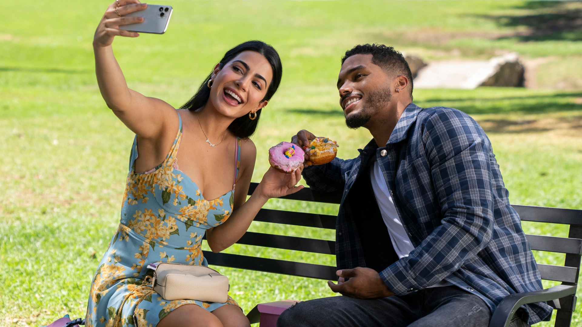Image from the show 'With Love' with two characters sitting on a bench taking a selfie while holding up donuts.