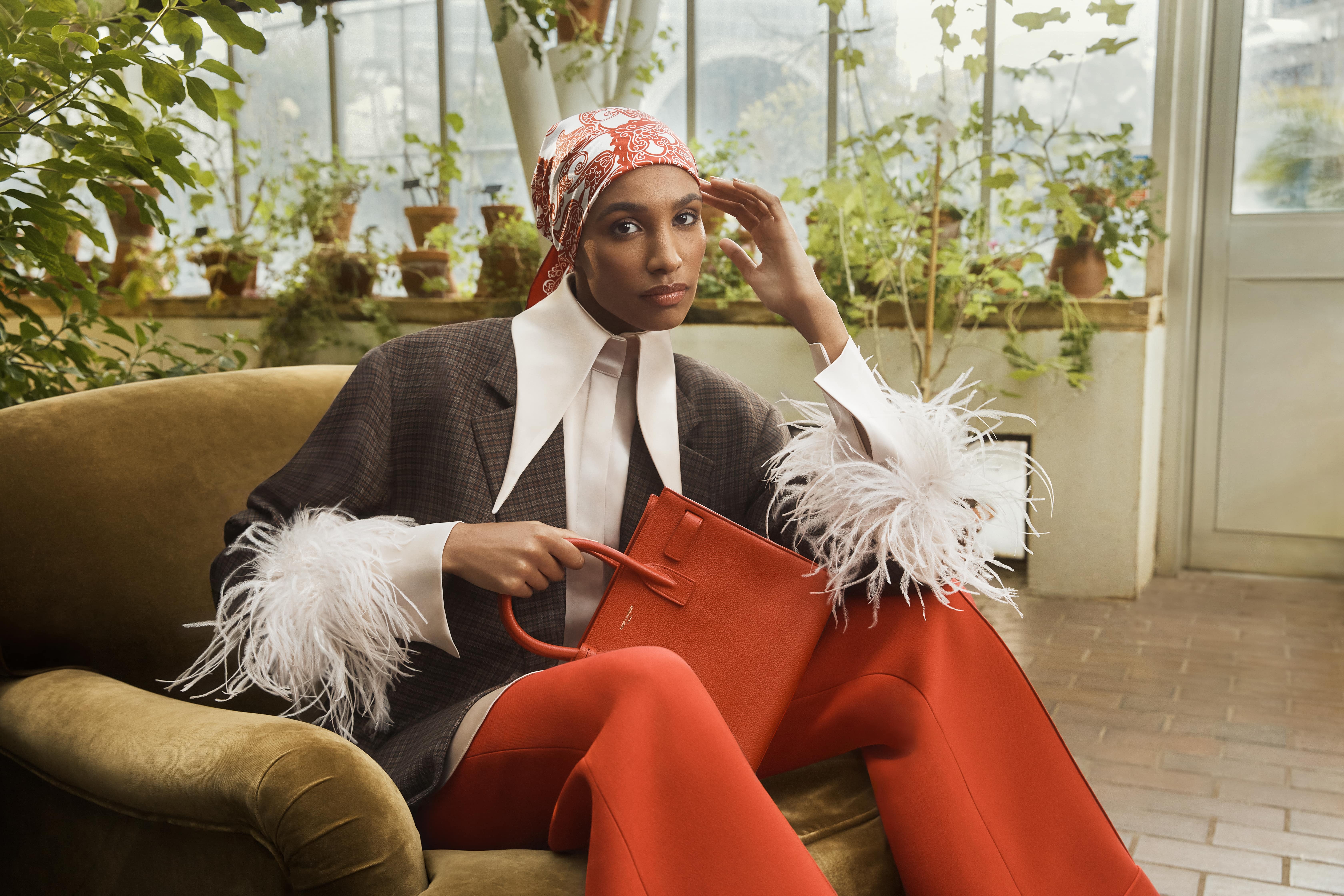 A woman on a single sofa in a lush green garden poses with an orange bag